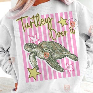 May include: White sweatshirt with a graphic of a sea turtle, stars, and the text "Turtley Over It" in green. The background features pink and white stripes. The turtle is green and brown.