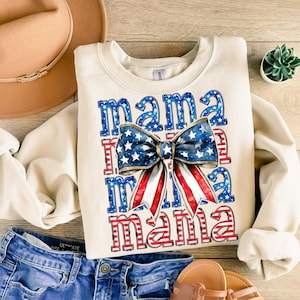 May include: Cream-colored sweatshirt with the word "mama" in red and blue, with a bow in the center featuring a stars and stripes design. The sweatshirt is paired with denim shorts, a tan hat, and sandals. A small succulent plant is also visible.