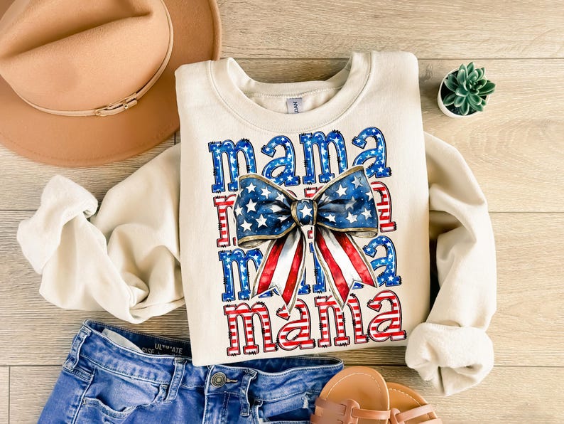 May include: Cream-colored sweatshirt with the word "mama" in red and blue, with a bow in the center featuring a stars and stripes design. The sweatshirt is paired with denim shorts, a tan hat, and sandals. A small succulent plant is also visible.