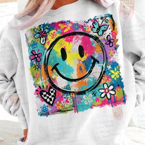 May include: White sweatshirt featuring a large, colorful graphic of a smiley face surrounded by flowers, butterflies, and a checkered heart. The design uses bright pink, yellow, blue, and green paint strokes for a vibrant, artistic look.