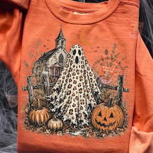 May include: An orange sweatshirt featuring a graphic print of a ghost with a leopard print pattern standing in front of a church and a wooden fence. Two pumpkins are in the foreground, and the text "WHAT A DESIGN" is visible in the upper right corner.