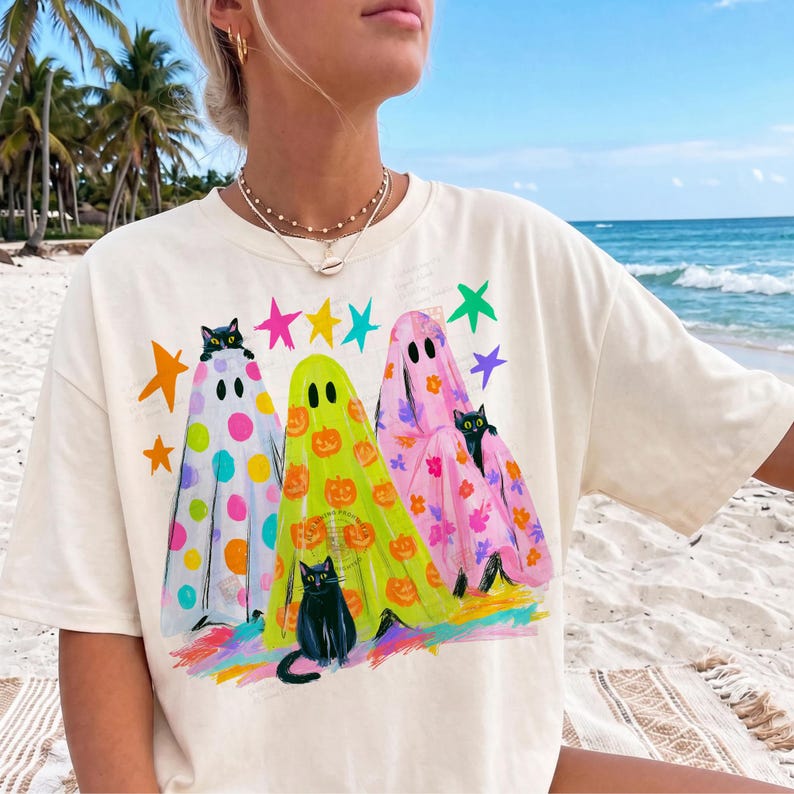 May include: A cream-colored t-shirt featuring a graphic print of three ghosts in white, lime green, and pink. The ghosts are decorated with colorful polka dots, orange pumpkins, and floral patterns, accompanied by black cats and bright stars.