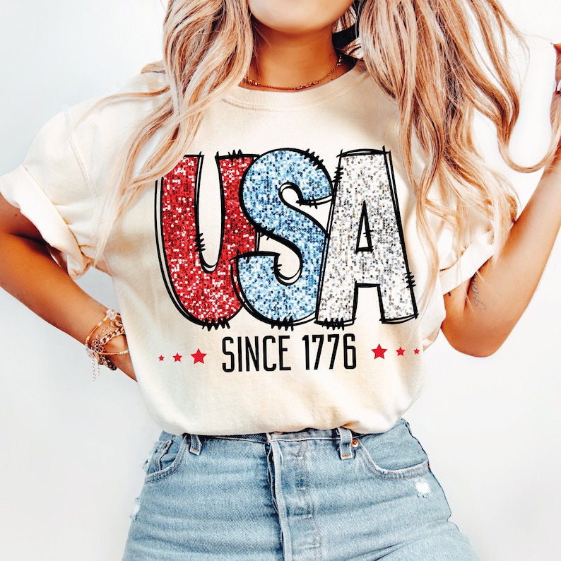 May include: A white t-shirt with a glittery red, white, and blue "USA" graphic and the text "SINCE 1776" underneath.