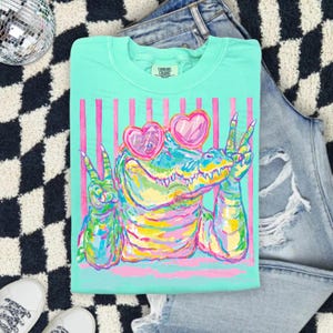 May include: A mint green Comfort Colors t-shirt featuring a colorful alligator wearing heart-shaped sunglasses and making peace signs. The design includes pink vertical stripes and the text "All Trading Rights".