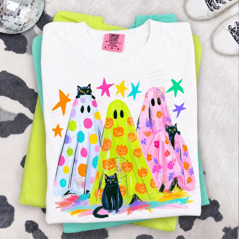 May include: A white t-shirt with a graphic print of three ghosts wearing patterned sheets, each accompanied by a black cat. The ghosts feature colorful polka dots, orange pumpkins, and floral designs, surrounded by bright neon stars and paint strokes.