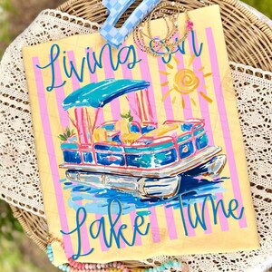 May include: A yellow tote bag with pink stripes and a painted pontoon boat design. The words "Living on Lake Time" are written in blue script. The boat is blue, pink, and white with a turquoise canopy and a yellow sun.