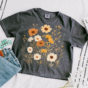 May include: A dark gray t-shirt with a floral print featuring yellow, orange, and white flowers. The flowers are arranged in a bouquet-like design.