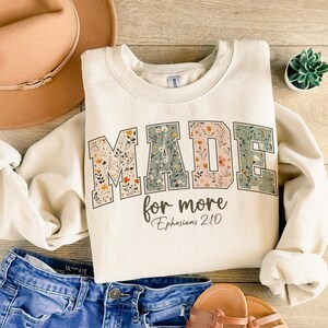 May include: Cream-colored sweatshirt with the words "MADE for more" in a floral design. The text "Ephesians 2:10" is below the main text. The sweatshirt is paired with blue denim shorts, a tan hat, and sandals. A small succulent plant is also visible.