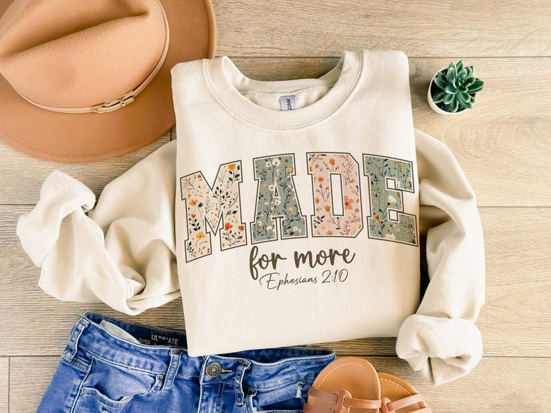 May include: Cream-colored sweatshirt with the words "MADE for more" in a floral design. The text "Ephesians 2:10" is below the main text. The sweatshirt is paired with blue denim shorts, a tan hat, and sandals. A small succulent plant is also visible.