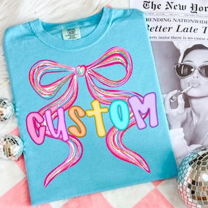 K&ouml;nnte beinhalten: Ein hellblaues T-Shirt mit einem gro&szlig;en, farbenfrohen Schleifendesign und dem Wort "CUSTOM" in bunten Buchstaben. Die Schleife hat rosa, goldene und blaue Akzente. Das Shirt liegt auf einer rosa-wei&szlig; karierten Oberfl&auml;che.