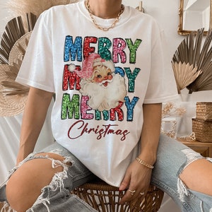 Op de afbeelding: Wit T-shirt met een kleurrijk glitterend "Merry Christmas" ontwerp met een vintage illustratie van de kerstman.