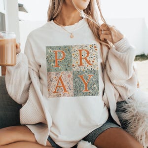 May include: A cream-colored crewneck sweatshirt featuring a square graphic divided into four quadrants. Each quadrant contains a different floral pattern in muted tones. The word PRAY is printed in large, bold, orange letters across the design.