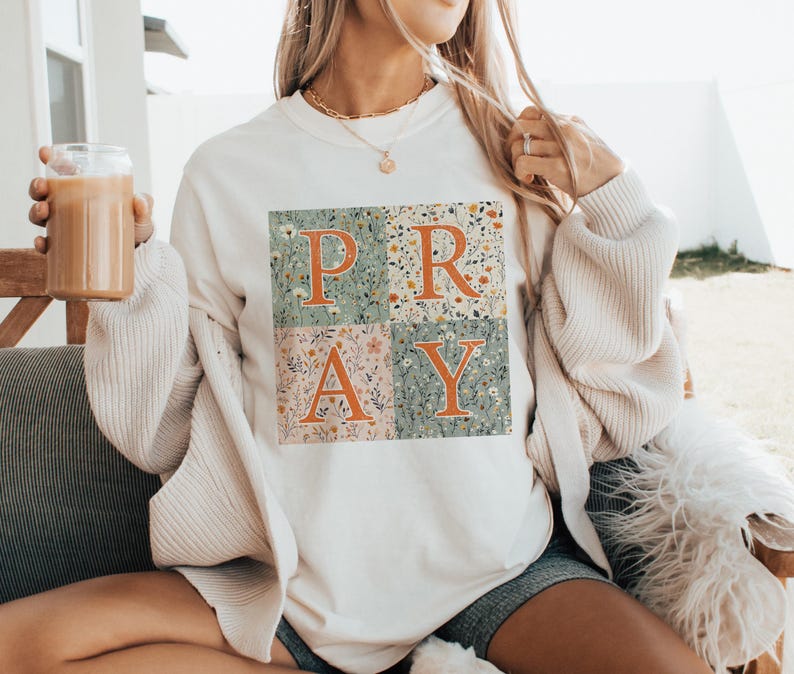May include: A cream-colored crewneck sweatshirt featuring a square graphic divided into four quadrants. Each quadrant contains a different floral pattern in muted tones. The word PRAY is printed in large, bold, orange letters across the design.