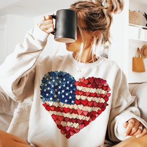 May include: White sweatshirt with a red, white, and blue heart-shaped American flag design. The flag is made of small hearts.