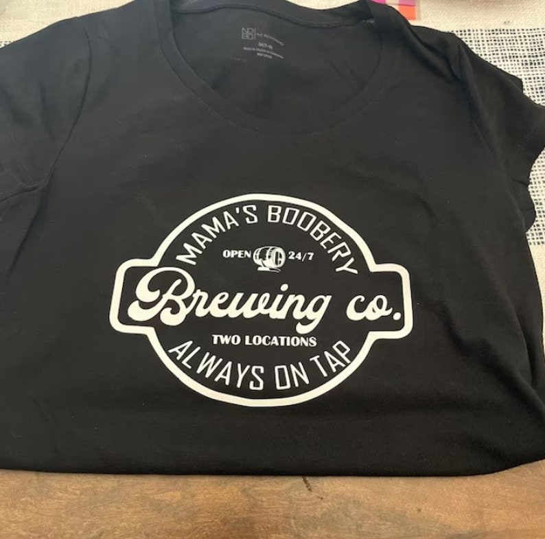 May include: Black t-shirt with a white circle logo that says "Mama's Booberry Brewing Co." with the text "Open 24/7" and "Two Locations Always On Tap" inside the circle.