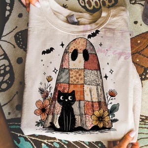 May include: A cream-colored sweatshirt featuring a patchwork ghost design with a black cat, flowers, bats, and stars. The ghost has a patchwork design in shades of orange, brown, and cream. The black cat sits at the bottom of the ghost.