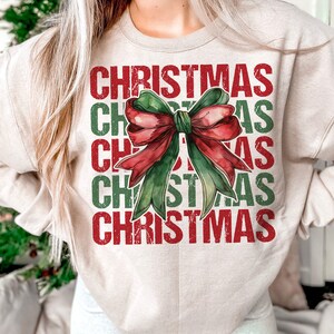 May include: Beige sweatshirt with the word "CHRISTMAS" in red and green block letters. A watercolor-style bow in red and green is centered on the front. The sweatshirt has long sleeves and a crew neck.