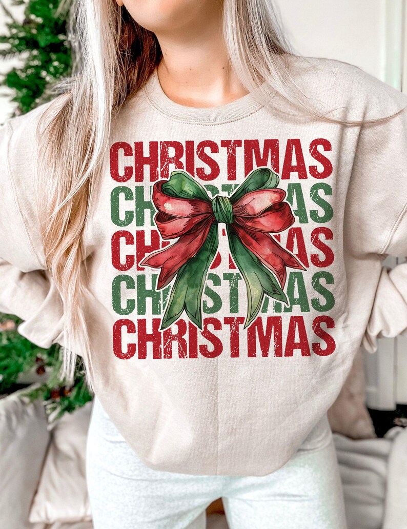 May include: Beige sweatshirt with the word "CHRISTMAS" in red and green block letters. A watercolor-style bow in red and green is centered on the front. The sweatshirt has long sleeves and a crew neck.