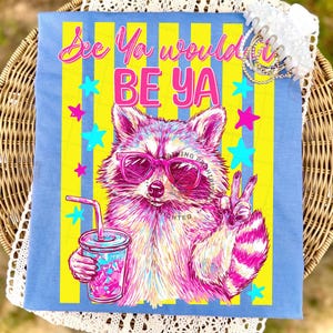 Peut inclure: T-shirt bleu clair avec un raton laveur rose et blanc portant des lunettes de soleil et tenant une boisson. Le t-shirt a des rayures jaunes et bleues et le texte "See Ya would BE Ya". Le raton laveur fait le signe de la paix.