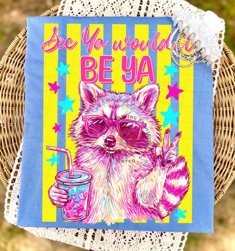 Peut inclure: T-shirt bleu clair avec un raton laveur rose et blanc portant des lunettes de soleil et tenant une boisson. Le t-shirt a des rayures jaunes et bleues et le texte "See Ya would BE Ya". Le raton laveur fait le signe de la paix.