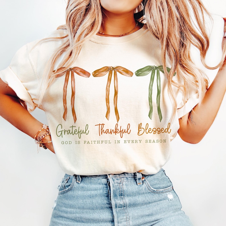 May include: Cream-colored t-shirt featuring three ribbon designs in brown, gold, and green. The words "Grateful, Thankful, Blessed" are printed below the ribbons in a fall color palette, with the text "GOD IS FAITHFUL IN EVERY SEASON" below.