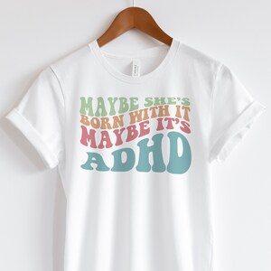 May include: White t-shirt with the text "MAYBE SHE'S BORN WITH IT MAYBE IT'S ADHD" in retro-style lettering. The text is in shades of green, orange, pink, and teal. The shirt is hanging on a wooden hanger.