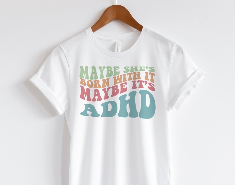 May include: White t-shirt with the text "MAYBE SHE'S BORN WITH IT MAYBE IT'S ADHD" in retro-style lettering. The text is in shades of green, orange, pink, and teal. The shirt is hanging on a wooden hanger.