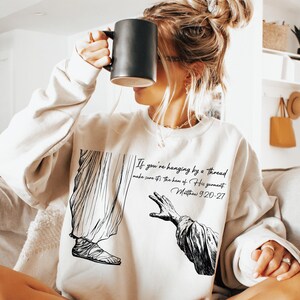 May include: A white sweatshirt featuring a black and white illustration of a hand reaching out from a robe. The text "If you're hanging by a thread make sure it's the hem of His garment. Matthew 9:20-27" is printed on the sweatshirt.