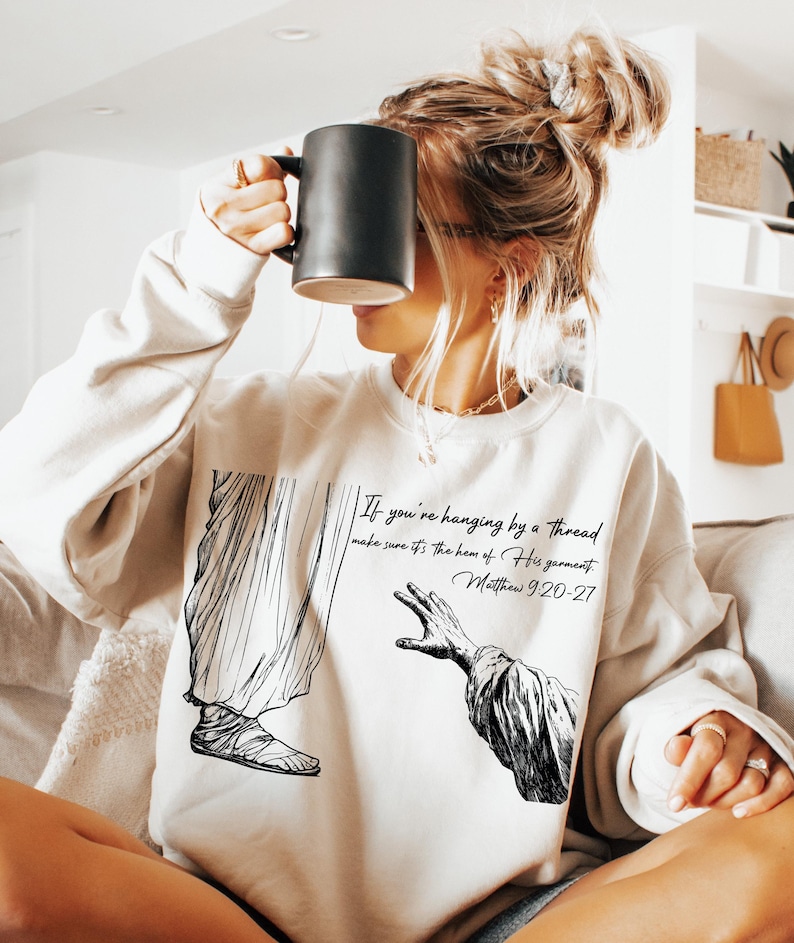May include: A white sweatshirt featuring a black and white illustration of a hand reaching out from a robe. The text "If you're hanging by a thread make sure it's the hem of His garment. Matthew 9:20-27" is printed on the sweatshirt.