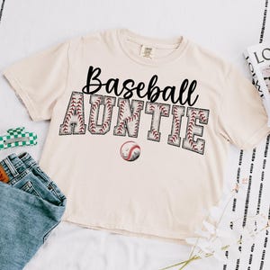 May include: A beige t-shirt with the text "Baseball Auntie" in black lettering. The text is surrounded by a red and white baseball stitch design. A baseball is pictured below the text.