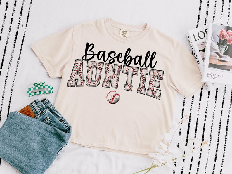 May include: A beige t-shirt with the text "Baseball Auntie" in black lettering. The text is surrounded by a red and white baseball stitch design. A baseball is pictured below the text.