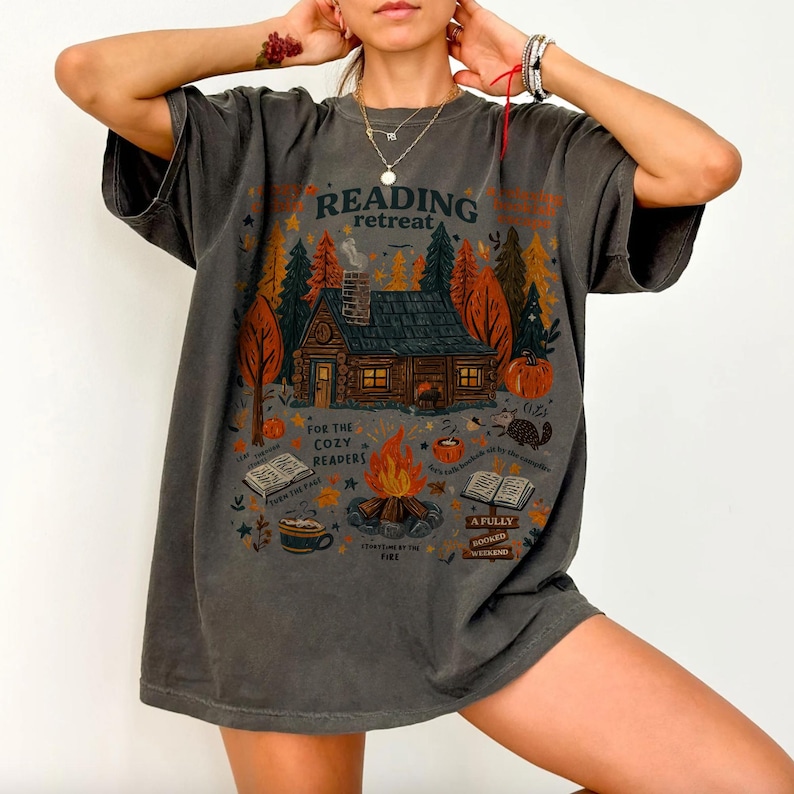 Pode incluir: Camiseta cinza escuro com um design gr&aacute;fico com uma cabana em uma floresta, uma fogueira e as palavras "Reading Retreat". O design inclui ab&oacute;boras, livros e a frase "For the Cozy Readers."