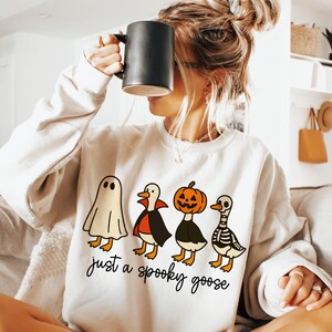 May include: Cream-colored sweatshirt featuring a Halloween-themed graphic of four cartoon geese dressed as a ghost, vampire, jack-o'-lantern, and skeleton. The text "just a spooky goose" is printed below the geese in a script font.