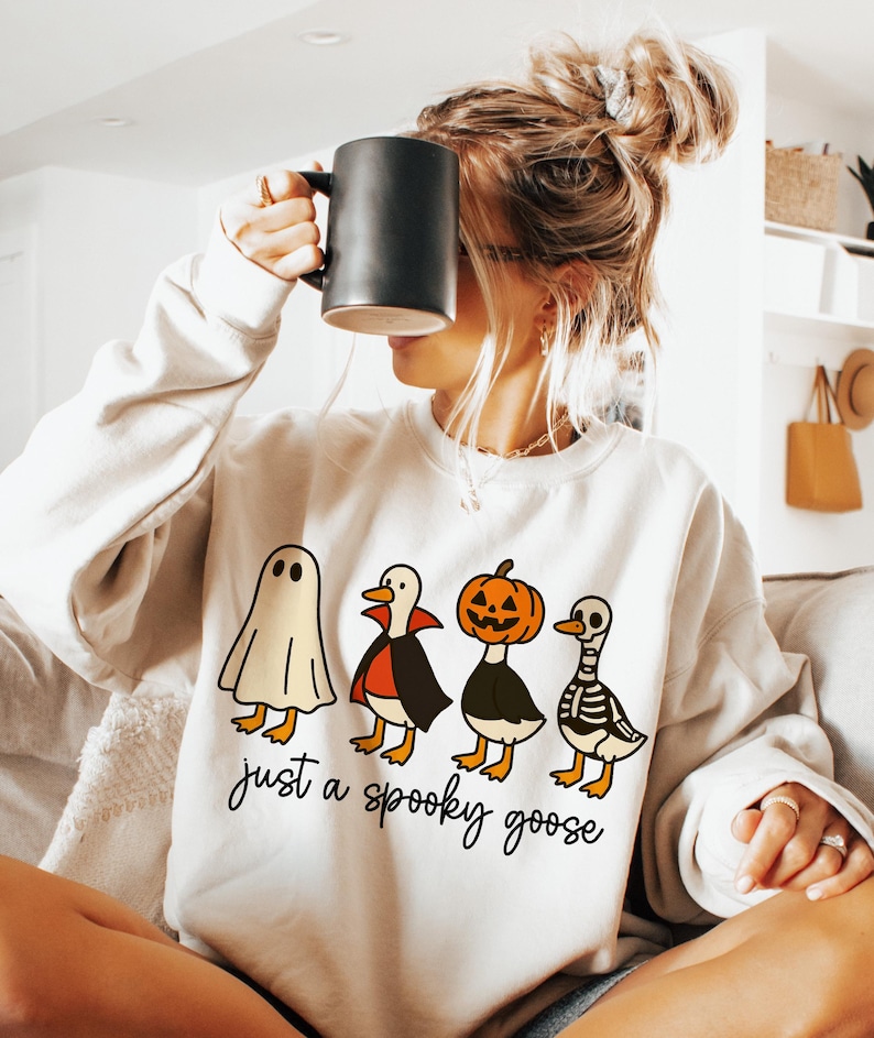 May include: Cream-colored sweatshirt featuring a Halloween-themed graphic of four cartoon geese dressed as a ghost, vampire, jack-o'-lantern, and skeleton. The text "just a spooky goose" is printed below the geese in a script font.