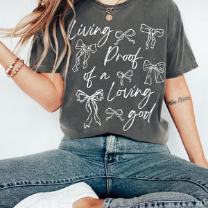 May include: A charcoal grey t-shirt with a white graphic design that says "Living Proof of a Loving Good" with a series of white bows around the text.