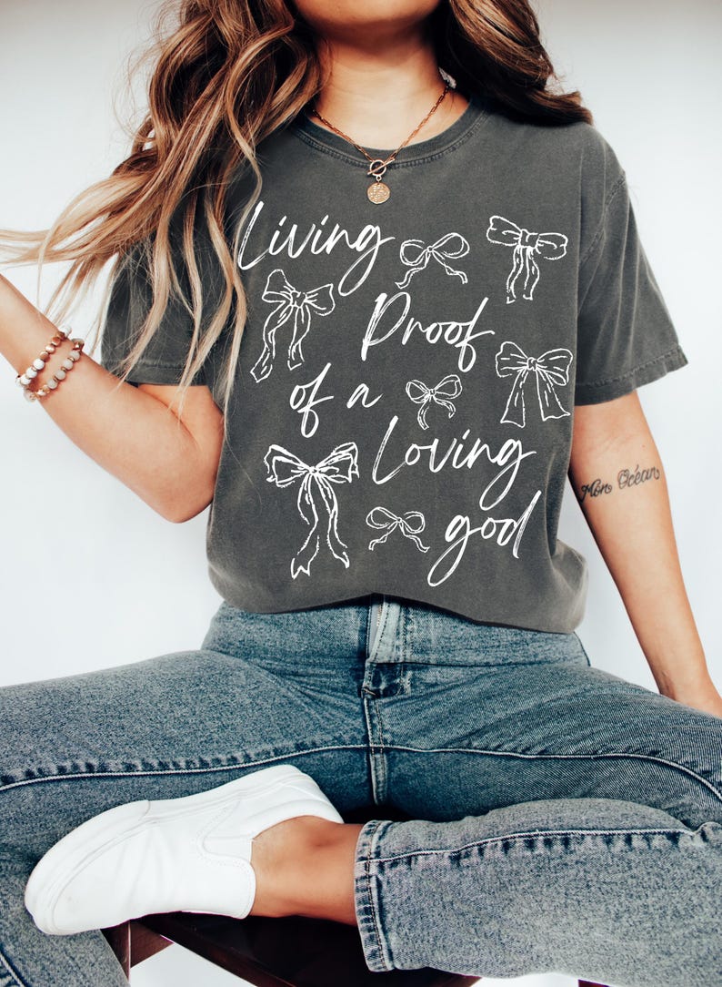 May include: A charcoal grey t-shirt with a white graphic design that says "Living Proof of a Loving Good" with a series of white bows around the text.
