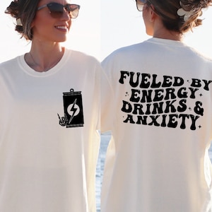 May include: White t-shirt with a black graphic of a can with a lightning bolt and a hand giving the rock and roll sign. The back of the shirt has the text "Fueled by Energy Drinks & Anxiety" in black.