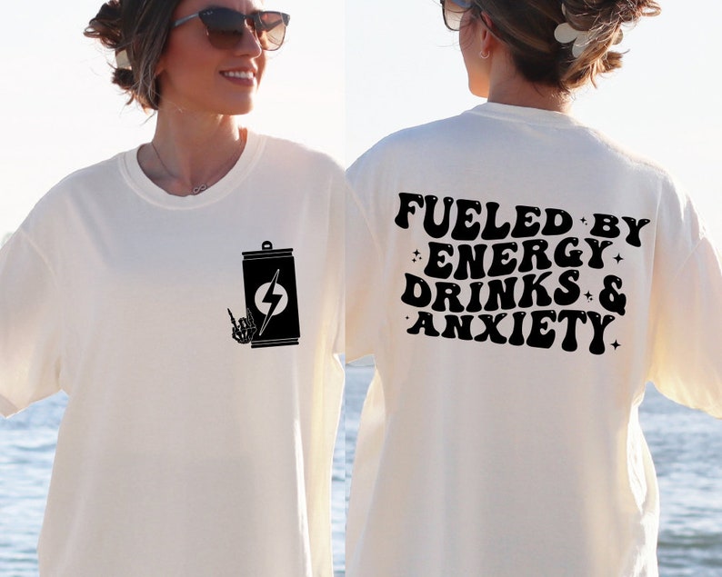 May include: White t-shirt with a black graphic of a can with a lightning bolt and a hand giving the rock and roll sign. The back of the shirt has the text "Fueled by Energy Drinks & Anxiety" in black.