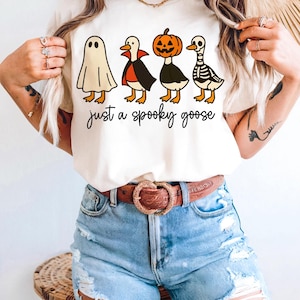 May include: Cream-colored t-shirt featuring four cartoon geese dressed in Halloween costumes: a ghost, a vampire, a jack-o'-lantern, and a skeleton. The text "just a spooky goose" is written in a black script font below the geese.
