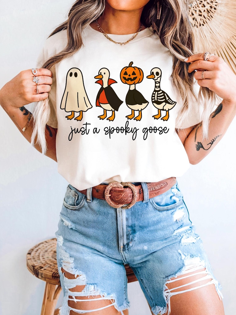 May include: Cream-colored t-shirt featuring four cartoon geese dressed in Halloween costumes: a ghost, a vampire, a jack-o'-lantern, and a skeleton. The text "just a spooky goose" is written in a black script font below the geese.
