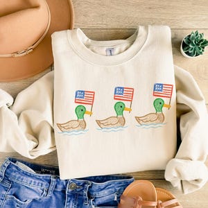May include: Cream-colored sweatshirt featuring an embroidered design of three ducks, each holding a small American flag. The ducks are depicted in green, brown, and yellow, with the flags in red, white, and blue. The sweatshirt is paired with blue denim shorts and a brown hat.