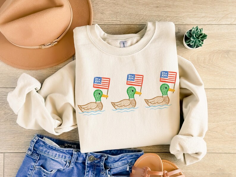 May include: Cream-colored sweatshirt featuring an embroidered design of three ducks, each holding a small American flag. The ducks are depicted in green, brown, and yellow, with the flags in red, white, and blue. The sweatshirt is paired with blue denim shorts and a brown hat.