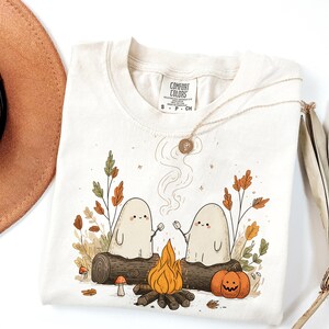 May include: Cream-colored Comfort Colors t-shirt featuring a campfire scene with two ghost characters roasting marshmallows. The design includes a bonfire, pumpkin, and fall foliage. A gold necklace and a brown hat are also visible.