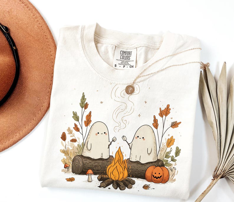 May include: Cream-colored Comfort Colors t-shirt featuring a campfire scene with two ghost characters roasting marshmallows. The design includes a bonfire, pumpkin, and fall foliage. A gold necklace and a brown hat are also visible.