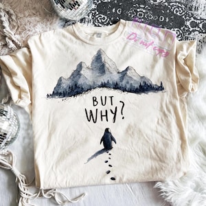 Könnte beinhalten: Cremefarbenes T-Shirt mit einer Illustration eines Bergmassivs in Grau- und Blautönen. Unter den Bergen steht der Text "BUT WHY?", darunter eine Pinguin-Illustration und Fußabdrücke.