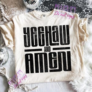 May include: Cream-colored t-shirt with the text "YEEHAW AND AMEN" in bold, black letters. The shirt is made of a soft material and has a classic crew neck. The shirt is displayed on a white fluffy surface.