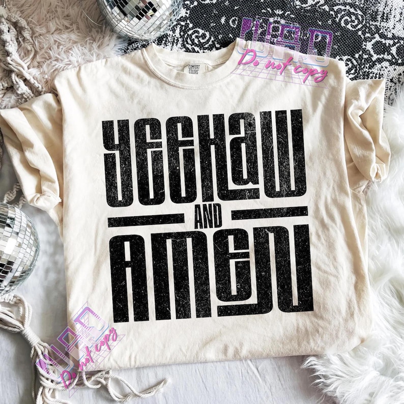 May include: Cream-colored t-shirt with the text "YEEHAW AND AMEN" in bold, black letters. The shirt is made of a soft material and has a classic crew neck. The shirt is displayed on a white fluffy surface.