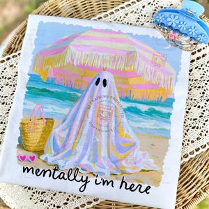 May include: A white t-shirt featuring a graphic of a ghost under a pink and yellow striped beach umbrella on a sandy beach. The design includes a straw tote bag, pink heart-shaped sunglasses, and the black text "mentally i'm here" at the bottom.