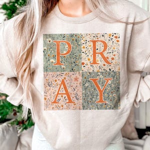 May include: A beige crewneck sweatshirt featuring a square graphic divided into four quadrants. Each quadrant displays a different floral pattern in muted tones. The orange letters P, R, A, and Y are overlaid across the floral background.