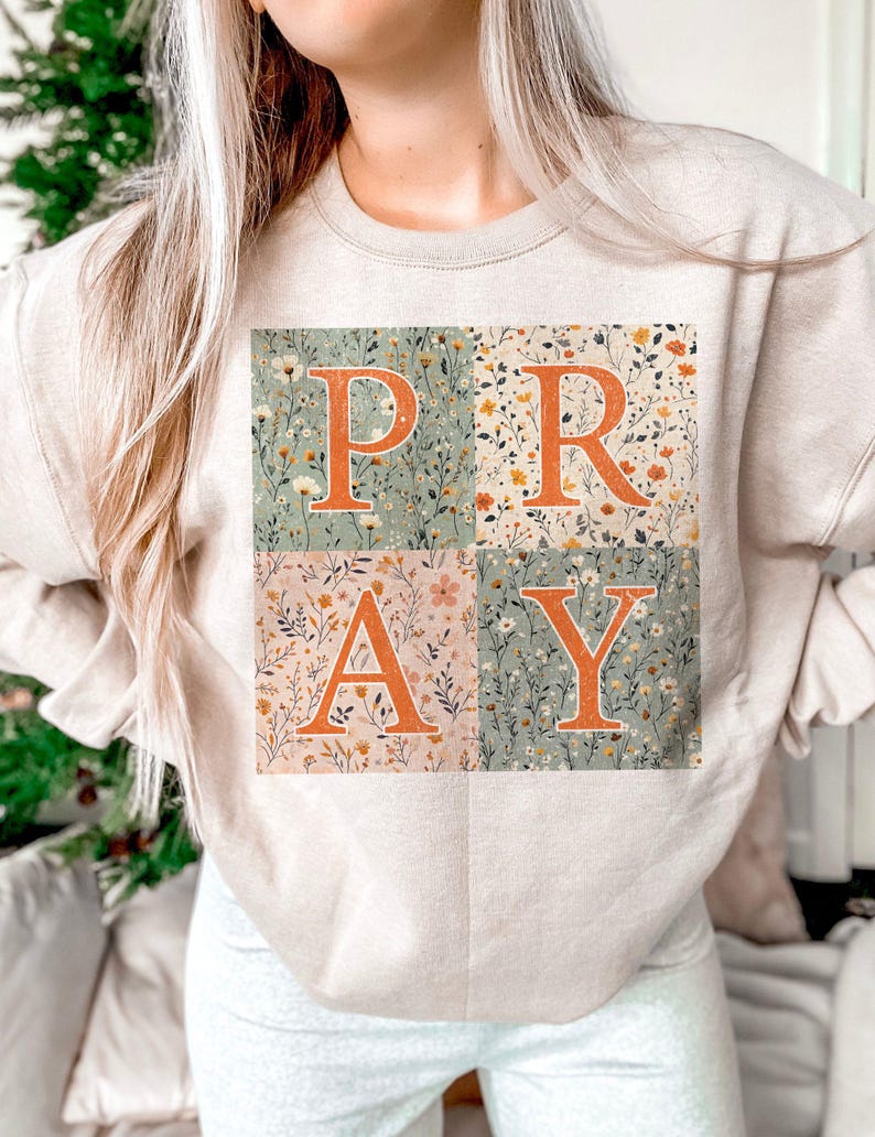 May include: A beige crewneck sweatshirt featuring a square graphic divided into four quadrants. Each quadrant displays a different floral pattern in muted tones. The orange letters P, R, A, and Y are overlaid across the floral background.